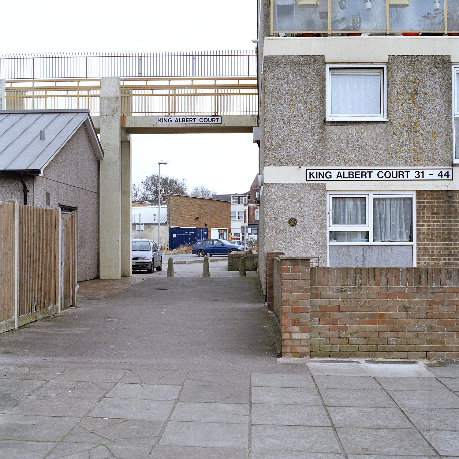 Pathway leads down the side of a block of flats and under an elevated walkway