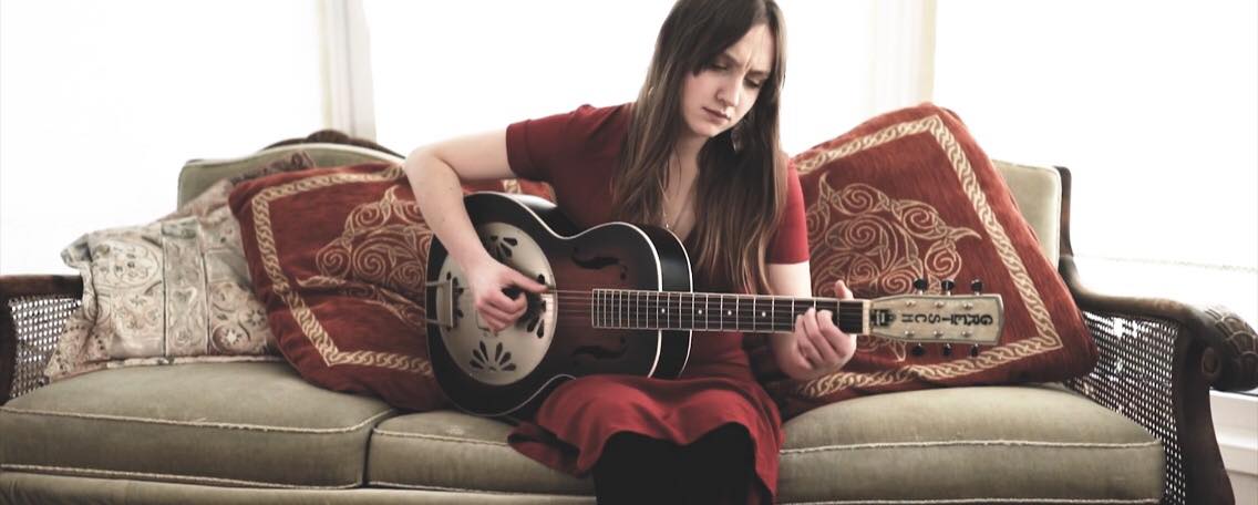 Megan Linford sat on a sofa playing her guitar. Photo by Tony Palmer