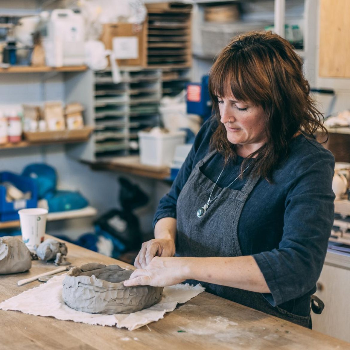 A woman with dark hair wearing a black shirt and an apron, working on a pottery piece